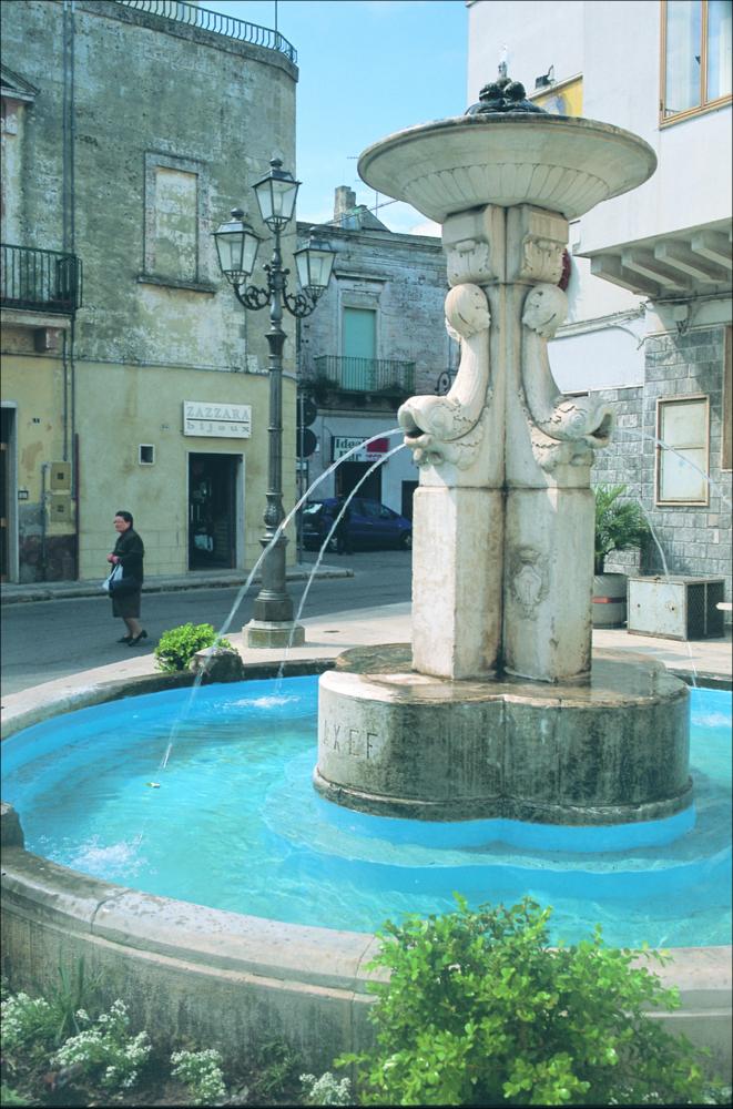 La fotografia è parte della ricca raccolta fotografica “Puglia per tutte le Stagioni” vol. 2, prodotta dall'Assessorato al Turismo e Industria Alberghiera della Regione Puglia, per raccontare la Puglia attraverso immagini suddivise in 5 settori principali: Città e Paesi, Settimana Santa, Territorio, Feste – Sagre – Tradizioni, Mercati – Prodotti – Cucina. La fotografia, scattata in originale a fine degli anni ‘70 mostra uno degli spazi urbani principali della città di Bari. Lo scatto immortala la fontana sita nel centro storico di Mottola, comune in provincia di Taranto. La particolarità del centro è quella di essere composto principalmente da case tinte di bianco, divenendo uno dei luoghi più belli e più importanti dal punto di vista turistico; oltre alla moltitudine d...
