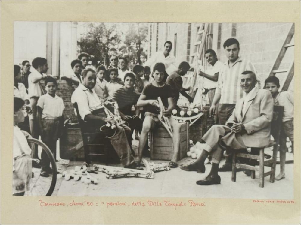 <div class="ql-editor"><div>Fotografia storica degli anni Cinquanta che ritrae i <strong>paratori</strong>, ovvero gli artigiani specializzati nella costruzione e nell’allestimento delle luminarie, della ditta Torquato Parisi. I soggetti, per lo più giovani affiancati da alcuni adulti, volgono lo sguardo verso l’obiettivo. L’immagine restituisce un raro momento di pausa e di consapevolezza, testimoniando la trasmissione intergenerazionale dei saperi artigianali e il ruolo centrale delle maestranze locali nella preparazione della Festa Patronale.</div><div><br></div><div>La <strong>Festa Patronale di Santo Stefano</strong> si celebra a Taurisano, in provincia di Lecce, nei giorni 2, 3 e 4 agosto. Sebbene non se ne conosca la data precisa di istituz...
