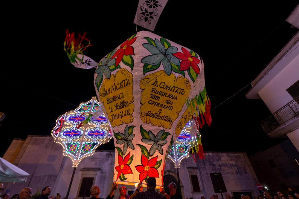 Si celebra la Festa Patronale di San Niceta a Melendugno e questo scatto fotografico immortala un momento di grande emozione per l’intera comunità: al rientro della processione, una folta folla di devoti assiste al lancio dei palloni aerostatici che portano su nel cielo preghiere e speranze.
La Festa di San Niceta Goto Martire, istituita nel 1740, si celebra nei giorni 15 e 16 Settembre a Melendugno in provincia di Lecce. Il rito religioso parte nove giorni prima della Festa, con delle celebrazioni eucaristiche presso la chiesa Madre e in forma esclusiva viene celebrata la santa messa in rito bizantino presso l’Abbazia di San Niceta situata in Melendugno sin dal 1167, riconosciuta appunto come uno dei più importanti riferimenti storici del territorio.
Il giorno della vi...