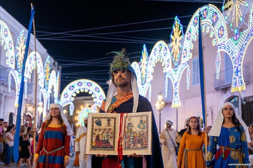 <div>La fotografia coglie in primo piano una comparsa del Corteo che regge un volume dalle pagine miniate, probabile riproduzione scenica di un codice medievale. Il libro, esibito come oggetto simbolico, richiama la trasmissione del sapere e la cultura scritta del tempo, mentre sullo sfondo il movimento del corteo accompagna la scena, inserendo il dettaglio in un più ampio racconto storico e rituale.</div><div><br></div><div>Il 13 agosto 2025, il <strong>Corteo Storico di Lucera </strong>ha animato le vie del centro, con partenza dalla Fortezza Svevo-Angioina e arrivo in piazza Duomo. La sfilata ha visto la partecipazione straordinaria di Sergio Muniz nelle vesti di re Carlo II d’Angiò e ha coinvolto oltre cinquecento figuranti – tra dame, cavalier...