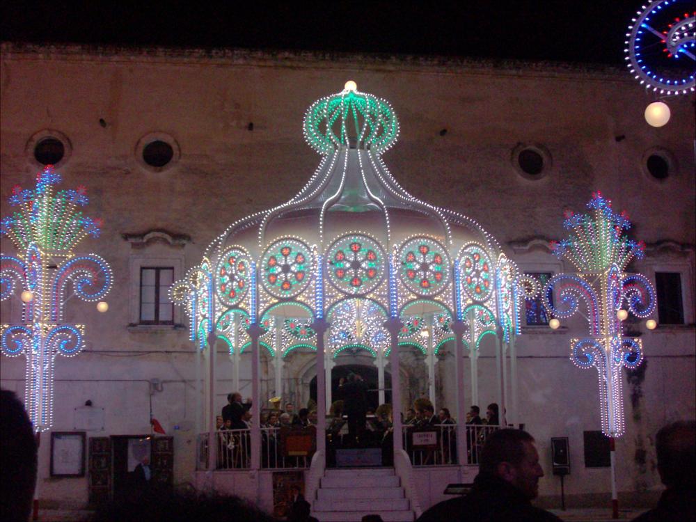 La fotografia mostra una cassa armonica illuminata durante la festa patronale di Montemesola, allestita come palcoscenico centrale per le esibizioni musicali. La struttura, decorata con luci e dettagli scenografici, ospita al suo interno il concerto della Banda Città di Montemesola “F. Trani”, diretta dal Maestro Beniamino Casavola, durante la celebrazione del 7 ottobre 2013.<div><br>La Festa Patronale della Beata Vergine del Santissimo Rosario di Montemesola (TA) è uno degli appuntamenti più sentiti e identitari della comunità. La celebrazione prende avvio nel fine settimana più vicino al 22 agosto, anniversario dell’elevazione della Vergine a Patrona del paese (22 agosto 1872), e si conclude il 7 ottobre, giorno della festività liturgica della Madonna del Rosario.</br...