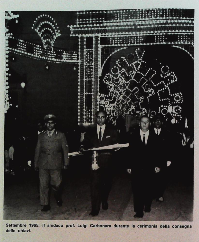La foto storica racconta un momento solenne del rito della consegna delle chiavi alla Santa Patrona della città di Triggiano da parte del sindaco prof. Luigi Carbonara nel settembre 1965. Sullo sfondo, luminarie e fedeli in processione fanno da cornice a questo importante e simbolico rito devozionale.
Le origini della devozione alla Madonna della Croce risalgono al XVI secolo, quando, fuori dall’antico abitato, venne edificata un’edicola sacra decorata con un’immagine “miracolosa” ad opera dei cosiddetti “homini de fora”, ossia contadini e braccianti che ogni giorno passavano di lì per andare a lavorare nei campi.
Una guarigione ritenuta miracolosa, avvenuta proprio nei pressi di quell’edicola, segnò l’inizio di una devozione destinata a crescere nel tempo, attirando se...