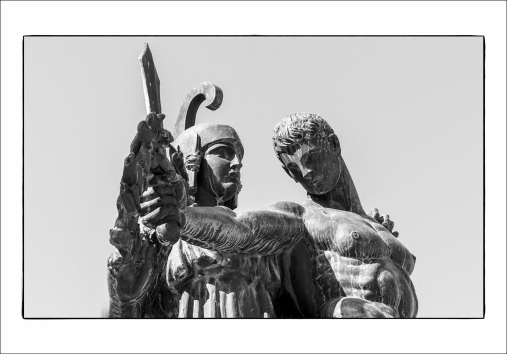 L'immagine digitale mostra un particolare del monumento ai Caduti a San Severo, opera dello scultore napoletano Amleto Cataldi formatosi tra la bottega del padre Angelo, intagliatore del legno, e la scuola romana di Ripetta. La scultura fu realizzata tra il 1918 ed il 1923 e fu posizionata al centro della odierna Piazza Luigi Allegato. Su un basamento quadrangolare si ergono due figure possenti alte circa due metri: una maschile, nuda secondo la tradizione greca, che solleva con la mano destra una spada e con la sinistra sorregge uno scudo simboli questi del combattente; alle spalle la figura femminile, personificazione della dea Minerva, con il capo cinto dall'Elmo di Scipio, allusione all'Italia, ha in una mano una ghirlanda d'alloro, destinata ai valorosi in battagli...
