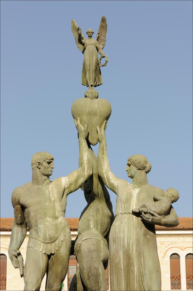 L'immagine digitale mostra un particolare del Monumento ai Caduti a Foggia, opera dello scultore napoletano Amleto Cataldi formatosi tra la bottega del padre Angelo, intagliatore del legno, e la scuola romana di Ripetta. La scultura risale al 1928 e fu posizionata al centro della odierna Piazza Italia. Protagonisti del dettaglio sono tre figure in bronzo, immortalate frontalmente, tre figure in bronzo: due eroi e una donna che sostiene un neonato, tutti reggono un cuore su cui è collocata una figuretta alata. I due eroi raffigurati con una possente muscolatura mostrano fieri il loro corpo: uno presenta un'armatura elegante, l'altro il corpo completamente nudo, fasciato sola da un perizoma. La figura maschile con l'armatura sostiene una spada e richiama il modello iconog...