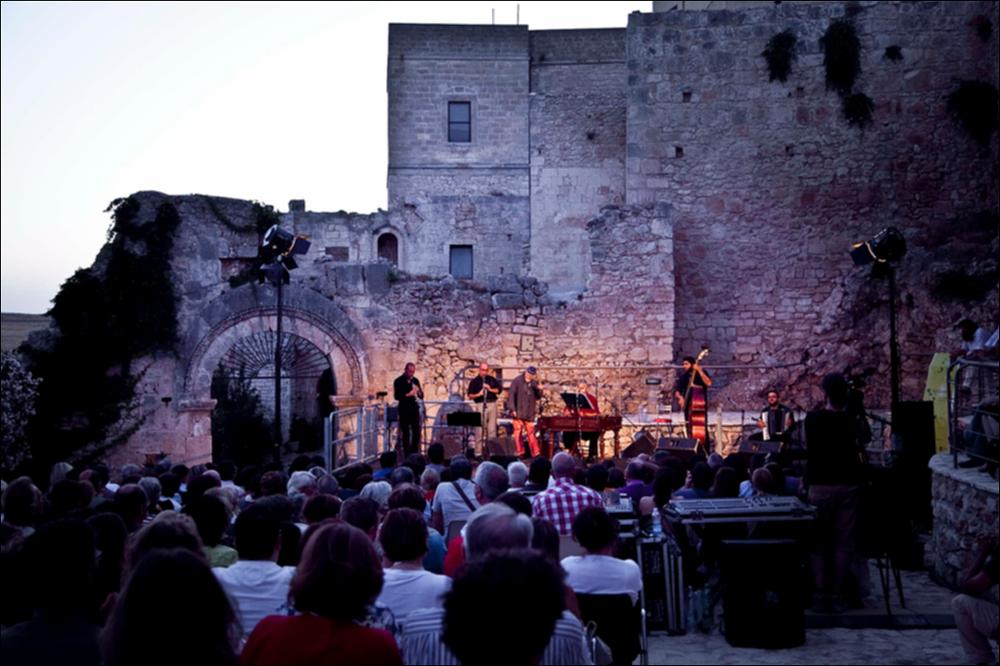 L'immagine digitale in oggetto è parte della collezione “Puglia Green Hour” nata dall'omonimo progetto di Teatro Pubblico Pugliese. Fu organizzato su un calendario fitto e intenso di spettacoli programmati tra giugno e luglio 2012 all'interno dei Parchi Regionali della Puglia, per parlare di arte, musica, teatro e filosofia ispirati dalle bellezze primordiali del paesaggio pugliese in aree territoriali spesso dimenticate o poco valorizzate. L'immagine, scattata poco dopo il tramonto, cattura un momento del concerto strumentale che si è tenuto l'8 luglio all'aperto, nell'area dell'Abbazia di Santa Maria di Pulsano, a Monte Sant'Angelo. L'antichissimo luogo sacro, simbolo di cristianità e spiritualità situato sul colle di Pulsano dal VI secolo, ha ispirato l'artista Moni ...