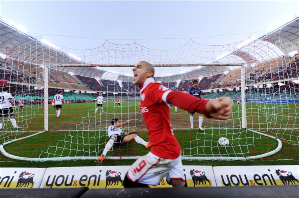 L'immagine digitale è uno scatto grandangolare del campo di calcio all'interno dello Stadio San Nicola di Bari durante una partita della squadra cittadina, scattata nel dicembre del 2014 in condizione di luce pomeridiana dal fotografo Sergio Scagliola. La fotografia cattura un momento di forte esultanza del giovane calciatore Giuseppe De Luca, in seguito ad un goal sferrato contro la squadra avversaria. Classe 1991, da luglio 2014 è attaccante del Bari, in prestito dall'Atalanta. Il grandangolare, eseguito dal bordo campo, mostra anche la struttura del maggior impianto sportivo di Puglia, lo Stadio San Nicola. Di proprietà del Comune di Bari, lo Stadio porta il nome del Santo Patrono barese, scelto a seguito di un insolito e trionfante referendum popolare effettuato tra...