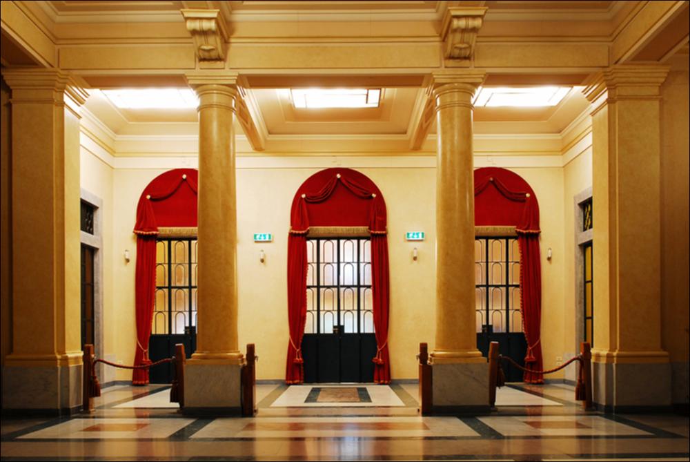 L'immagine è parte della campagna fotografica realizzata dalla fotografa Margherita Mirabella per Teatro Pubblico Pugliese che, con i suoi scatti, ha ripreso le eleganti architetture e i rifiniti dettagli di alcuni dei più bei teatri pugliesi e di alcuni spettacoli teatrali portati in scena in questi ultimi anni. La fotografia ci restituisce il foyer del Teatro Comunale Giuseppe Verdi di San Severo riconosciuto come la più grande struttura teatrale di Capitanata e terzo teatro all'italiana più importante di Puglia, dopo il Petruzzelli di Bari e il Politeama Greco di Lecce. Il monumento, originariamente denominato teatro Comunale del Littorio, viene progettato dall'architetto Cesare Bazzani rispettando a pieno i canoni stilistici dell'arte fascista. I lavori, iniziati ne...