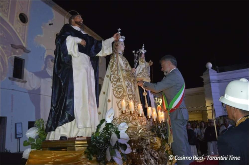 La fotografia ritrae un momento centrale della festa patronale: la consegna simbolica delle chiavi della città da parte del sindaco alla Madonna del Rosario, posta al centro della scena, affiancata dai simulacri di San Domenico e Santa Caterina. Il sindaco, in abito formale, porge le chiavi verso il simulacro centrale, gesto che simboleggia l’affidamento della città alla Vergine. <div><br>La Festa Patronale della Beata Vergine del Santissimo Rosario di Montemesola (TA) è uno degli appuntamenti più sentiti e identitari della comunità. La celebrazione prende avvio nel fine settimana più vicino al 22 agosto, anniversario dell’elevazione della Vergine a Patrona del paese (22 agosto 1872), e si conclude il 7 ottobre, giorno della festività liturgica della Madonna del Rosario...