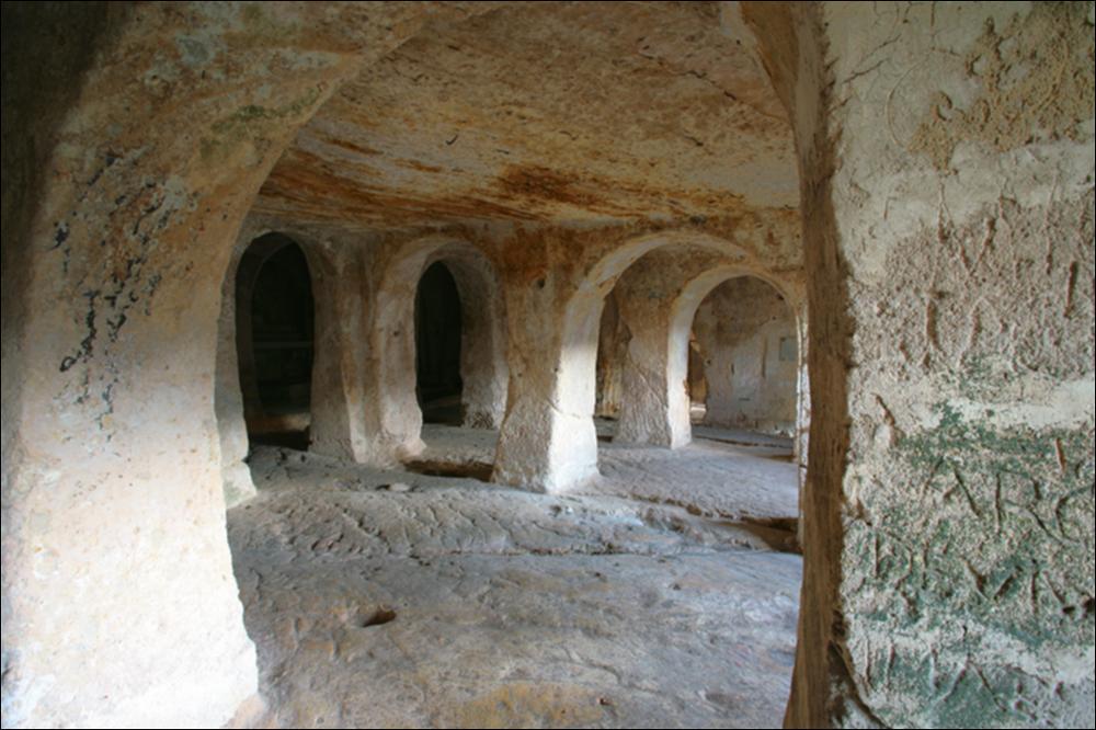 L'immagine digitale rappresenta un particolare, immortalato dal fotografo pugliese Carlos Solito, dell'interno della chiesa-grotta di San Michele, sita in Gravina in Puglia. La chiesa, circondata da grotte più piccole, si trova nel rione Fondovico, in un complesso rupestre distribuito su tre superfici non più esistenti. Scavata in un unico masso di tufo granulare, è sorretta da 14 pilastri naturali di forma quadrangolare che dividono la chiesa in cinque navate. Al suo interno è possibile osservare parti di affreschi, tra cui un Cristo Pantocratore tra San Michele e San Paolo (secc. XII-XIV) e tre altari con le rispettive statue in pietra e gesso, seicentesche e settecentesche, degli arcangeli Michele, Gabriele e Raffaele, oltre una tomba medievale ad arcosolio e il dipi...