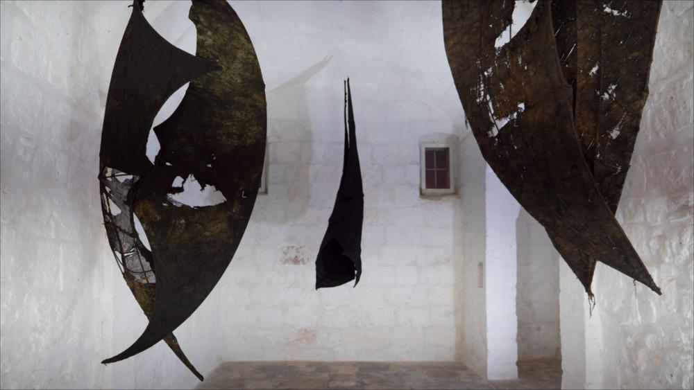 L’immagine presenta Lazarus 2021, una delle opere di Ibrahim Mahami, vincitore del Premio Pascali 2021 (XXIII edizione), in mostra presso la Fondazione Museo Pino Pascali di Polignano a mare dall’11 dicembre 2021 al 13 marzo 2022.
L’installazione è stata collocata nella "exchiesetta", chiesa sconsacrata di Santo Stefano, e si tratta di un’altra opera ispirata ai numerosi sili costruiti negli anni Sessanta a Tamale.
L’installazione si compone di una serie di sculture, formate da armature di tondini metallici, avvolte dai teloni usati per coprire i carichi durante le spedizioni internazionali. Le tele cerate, impregnate di olio a causa del loro continuo contatto con le parti meccaniche dei mezzi di trasporto, si trasformano nelle figure ammantate dei pipistrelli che, sosp...