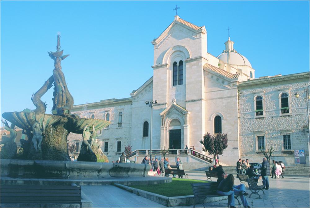La fotografia è parte della ricca raccolta fotografica “Puglia per tutte le Stagioni” vol. 2, prodotta dall'Assessorato al Turismo e Industria Alberghiera della Regione Puglia, per raccontare la Puglia attraverso immagini suddivise in 5 settori principali: Città e Paesi, Settimana Santa, Territorio, Feste – Sagre – Tradizioni, Mercati – Prodotti – Cucina. L'immagine raffigura Piazza Vittorio Emanuele II a Giovinazzo, in una bella giornata di sole. La piazza, ampio e raffinato salotto cittadino, ha una caratteristica forma trapezoidale, al centro si scorge la fontana monumentale dei Tritoni realizzata in bronzo e in pietra locale su progetto dello scultore giovinazzese Tommaso Piscitelli e inaugurata nel 1933. Ma le origini della piazza risalgono al 1797, quando fu costr...