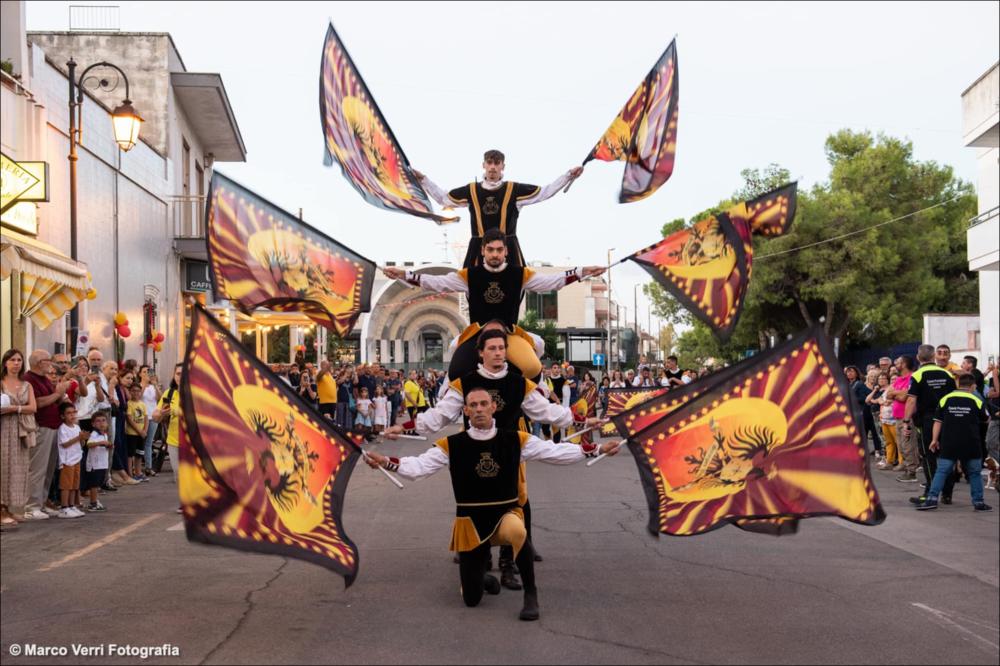In questo scatto del 2022 sono protagonisti un gruppo di sbandieratori che animano le vie cittadine di Melendugno in occasione della Festa Patronale di San Niceta.
La Festa Patronale di San Niceta Goto Martire, istituita nel 1740, si celebra nei giorni 15 e 16 Settembre a Melendugno in provincia di Lecce. Il rito religioso parte nove giorni prima della Festa, con delle celebrazioni eucaristiche presso la chiesa Madre e in forma esclusiva viene celebrata la santa messa in rito bizantino presso l’Abbazia di San Niceta situata in Melendugno sin dal 1167, riconosciuta appunto come uno dei più importanti riferimenti storici del territorio.
Il giorno della vigilia della Festa, come da tradizione pluricentenaria, al termine della Santa Messa, i fedeli accompagnati da un concer...