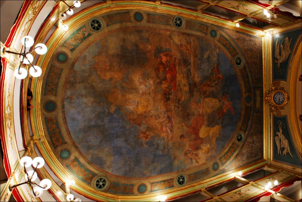 L'immagine è parte della campagna fotografica realizzata dalla fotografa Margherita Mirabella per Teatro Pubblico Pugliese, che con i suoi scatti ha ripreso le eleganti architetture e i rifiniti dettagli di alcuni dei più bei teatri pugliesi e di alcuni spettacoli teatrali portati in scena in questi ultimi anni. La fotografia ritrae l'affresco che domina e decora il soffitto del Teatro Paisiello di Lecce. Il dipinto fu realizzato dal pittore napoletano Vincenzo Paliotti quando, nel terzultimo decennio dell'800, il teatro viene ricostruito in pietra e completato con delicati elementi decorativi affidati a maestranze napoletane. Il maestro Palmiotti alle figure delle Grazie accosta le allegorie della Commedia, della Tragedia e dell'Armonia. Nel 1870 il teatro, dedicato al...